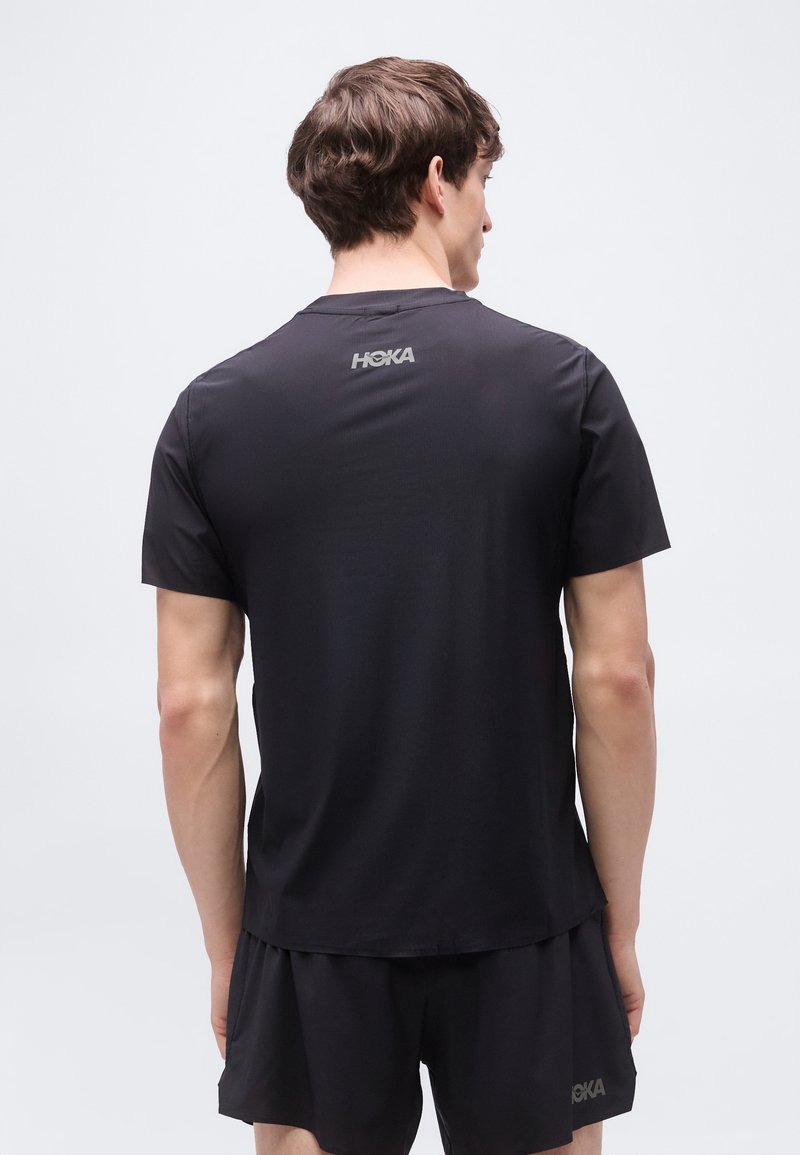 Uomo di spalle che indossa una maglietta sportiva nera HOKA e pantaloncini abbinati su uno sfondo chiaro e uniforme.