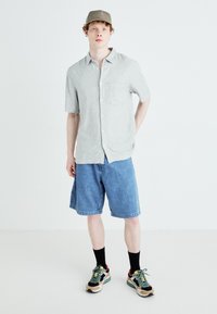 Chemise boutonnière à manches courtes gris clair avec une poche poitrine, associée à un short en denim bleu et des baskets colorées. Inclut des chaussettes noires à la cheville.