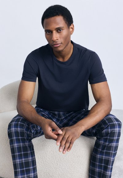 Tamsiai mėlyna marškinėliai su apvalia iškirpte derinami su languotomis pižamos kelnėmis, kuriose dominuoja mėlynos, juodos ir baltos spalvos; žmogus sėdi ant tekstūruoto, šviesios spalvos paviršiaus.