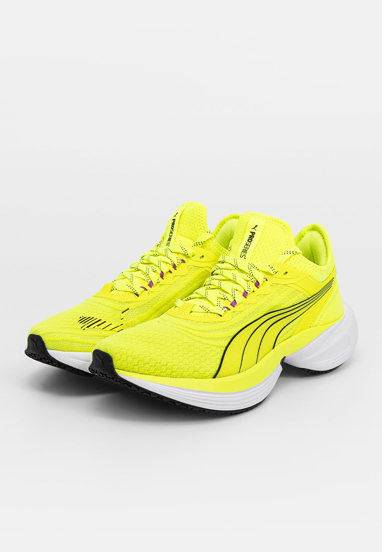 Neon żółte buty do biegania z teksturowaną siateczką, czarnymi akcentami i fioletowymi szczegółami sznurówek. Posiadają wyściełaną białą podeszwę dla wsparcia.