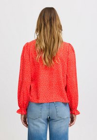 Röd blus med vita prickar, långa puffärmar och volangärmar. Mjuk tyg med avslappnad passform, kombinerad med ljusblå jeans.