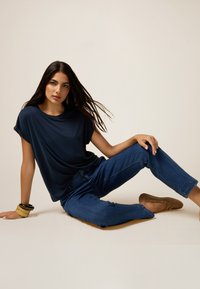 Navigatieblauwe, losse t-shirt met opgerolde mouwen, gecombineerd met nauwsluitende donkere spijkerbroek. Model draagt bruine instapschoenen en gelaagde armbanden.
