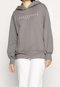 Szary hoodie z mieszanki bawełny z przednią kieszenią typu kangur, ściągaczami przy mankietach i dolnej krawędzi oraz wytłoczonym logo "Abercrombie New York" na klatce piersiowej.