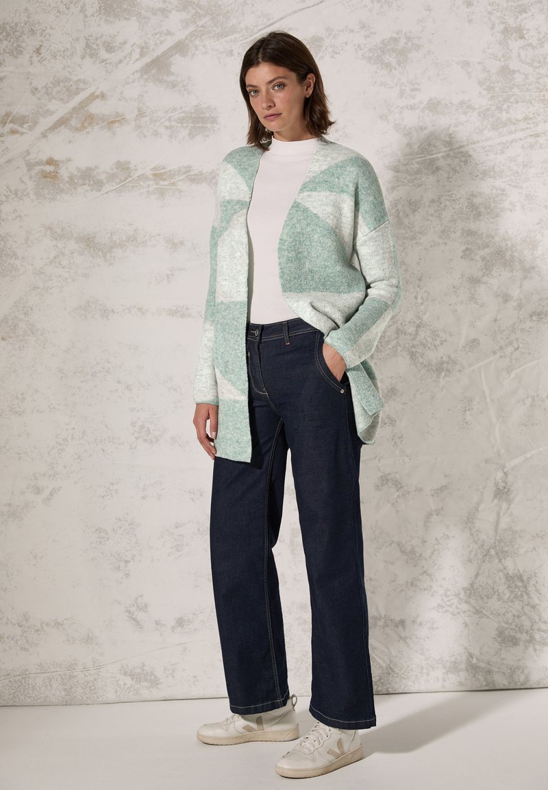 Licht mintgroene cardigan met een textuurpatroon, gedragen over een witte top; gecombineerd met donkerblauwe jeans en witte sneakers.