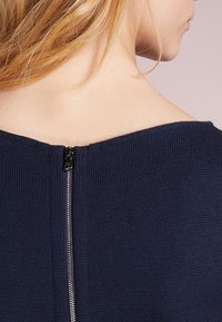 Mörkblå stickad topp med bred halsringning, med en silverfärgad dragkedja bak. Tyget ser strukturerat och åtsittande ut.