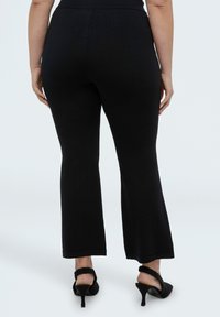 Mujer vistiendo pantalones negros ajustados y recortados con campana y zapatos slingback negros de tacón bajo, de pie contra un fondo blanco.