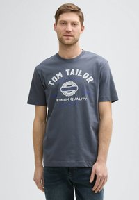 Camiseta de algodón gris con el texto "TOM TAILOR" en blanco y azul junto a un gráfico de insignia circular. Mangas cortas, cuello redondo, corte relajado.