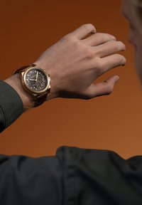 Orologio cronografo con cassa in bronzo, quadrante nero e cinturino in pelle marrone. Presenta più sotto-quadranti e una finitura texturizzata.