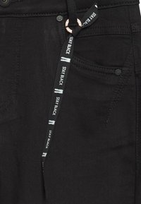 Blugi negri din denim, cu șnur pe care este inscripționat textul „STAY BLACK”, nasture din cupru și design cu cinci buzunare. Textură netedă, croială mulată.