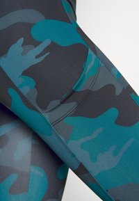 Legíny s kamuflážním vzorem v odstínech tyrkysové a tmavě šedé. Vyrobené z elastického materiálu se hladkým povrchem a plochými švy.