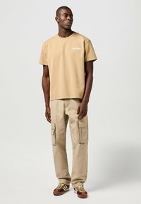 Beige bomulls-t-shirt med kort ärm och "Wrangler"-logotyp, ihop med beige cargobyxor med två sidofickor och bruna sneakers.