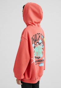 Sudadera coral de gran tamaño con un bolsillo canguro delantero. El diseño gráfico en la parte posterior presenta elementos coloridos y texto, fabricada en suave material de algodón.