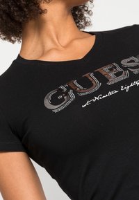 Schwarzes V-Ausschnitt-T-Shirt mit einem silbernen Strass-Logo "GUESS" und geschwungener Schrift, taillierte Passform mit kurzen Ärmeln und weichem Baumwollmaterial.