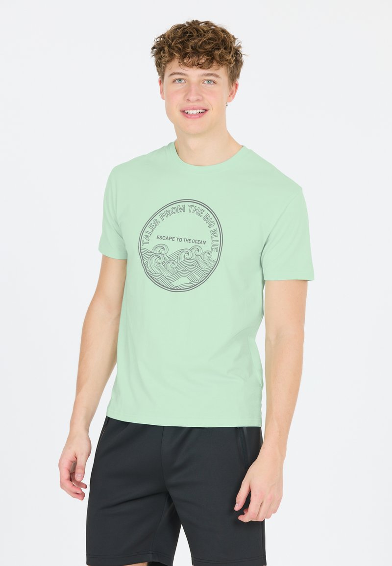 Ljusgrön t-shirt med kort ärm som har en cirkulär grafik med vågor och text, kompletterad med svarta sportshorts. Slät tygtextur.