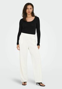 Schwarzes, geripptes Langarmoberteil mit gewelltem Kragen, kombiniert mit weißen, weiten Hosen. Das Outfit zeichnet sich durch eine glatte Textur und minimale Akzente aus.