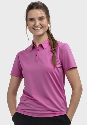 Lächelnde Frau mit langem geflochtenem Haar, die ein pinkes kurzärmeliges Poloshirt und schwarze Hosen trägt, steht mit den Händen in den Taschen.