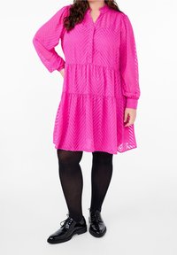 Růžové šaty s dlouhým rukávem a zapínáním na knoflíky, se zvýrazněným volánkovým designem a chevron vzorem. Měly na sobě černé lakové boty a punčochy.