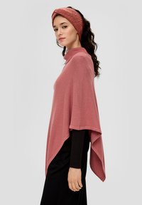 Roze gebreide poncho met een hoge kraag, textuurstof en een asymmetrische zoom, gecombineerd met een bijpassend haarbandje. Slanke, zwarte top eronder.
