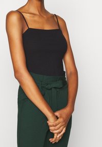 Black ribbed tank top with thin straps, paired with high-waisted green trousers featuring a bow tie at the waist. Smooth texture.
