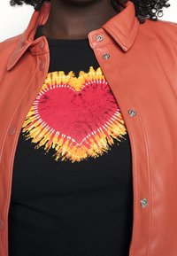 Giacca di pelle arancione con bottoni a pressione indossata sopra una t-shirt nera con un disegno di cuore rosso e giallo dai bordi appuntiti.
