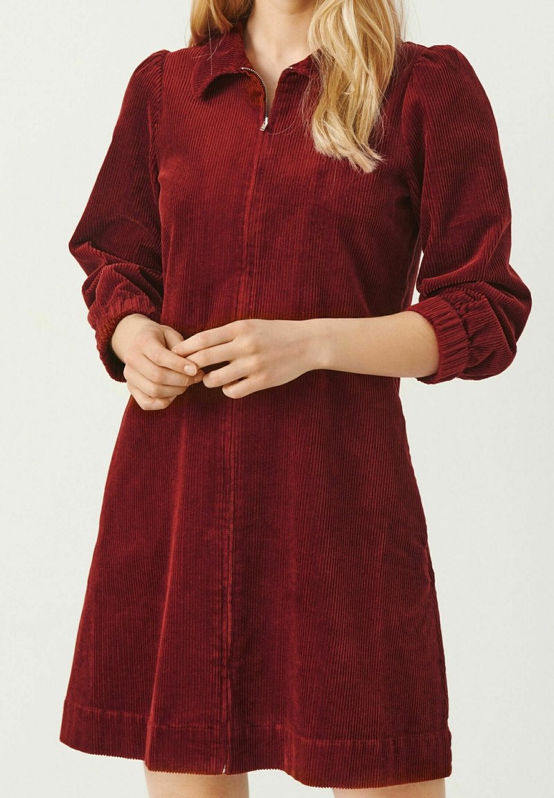 Robe en velours rouge avec un col et une fermeture éclair à l'avant. Présente de longues manches bouffantes avec des poignets élastiques et une jupe évasée. Surface texturée.