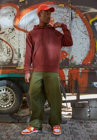 Mörkröd huvtröja med en framficka, lösa olivgröna byxor och flerskiktade sneakers med orangea och vita detaljer, mot en bakgrund av graffiti.