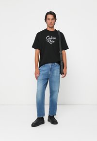 Camiseta de algodón negra con logo blanco de "Calvin Klein", corte holgado. Jeans de mezclilla azul claro, corte recto, con dobladillo en la parte inferior, combinados con zapatillas negras.