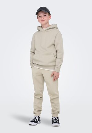 Ragazzo in piedi con le mani nelle tasche, indossa una felpa con cappuccio beige, pantaloni della tuta abbinati, un berretto nero e sneakers alte nere su sfondo semplice.