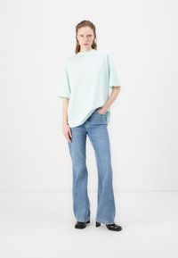 Mátyově zelené oversized tričko s vysokým výstřihem, spárované s světle modrými rozšířenými džíny. Model stojí na bílém pozadí.