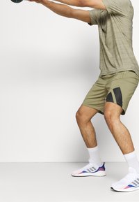 Urheiluasu, jossa on vaaleanvihreä lyhythihainen paita ja khakiväriset shortsit, joissa on musta viistovivahde. Valkoiset urheilukengät, joissa on monivärinen pohja.