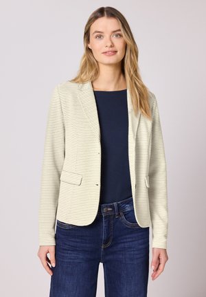 Giovane donna che indossa un blazer crema testurizzato sopra una maglietta blu navy e jeans blu scuro, in piedi davanti a uno sfondo chiaro e uniforme.