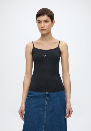 Γυναίκα με κοντά μαλλιά που φοράει εφαρμοστό μαύρο τοπ με μικρό λευκό λογότυπο και μπλε τζιν φούστα, στέκεται μπροστά σε απλό φόντο.