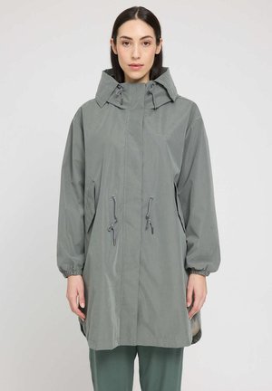 Woman with straight dark hair wearing a light gray hooded long raincoat with drawstrings, standing against a plain light background.