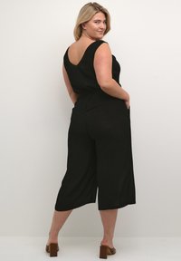 Svart linne jumpsuit med vida byxor, V-ryggdesign och sidofickor. Bärs med bruna blockklack-sandaler.