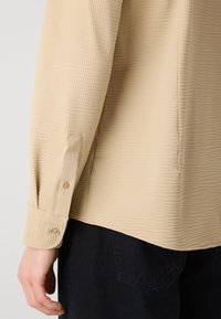 Camisa texturizada beige con puños de botón y un ajuste relajado, que presenta un patrón tejido detallado, combinada con pantalones de tela oscuros.