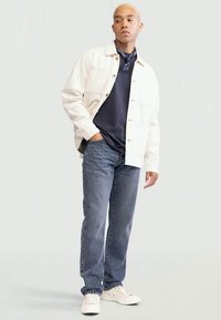 Casaco branco, t-shirt azul-marinho, jeans azuis, ténis brancos. Apresenta frente com botões, bolsos laterais e um lenço estampado ao redor do pescoço.