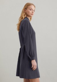 Vestito grigio scuro a maniche lunghe con colletto alla cinese, polsini elasticizzati e cintura in vita, caratterizzato da una silhouette rilassata e una texture morbida.