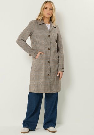 Blonde vrouw met een geruite jas tot op de knie over een wit shirt, blauwe wijde jeans en witte sneakers, staand tegen een effen achtergrond.