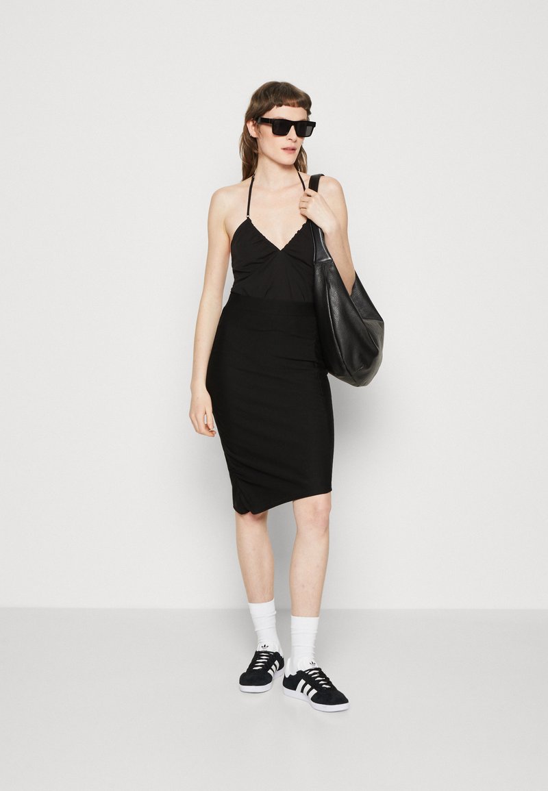 Schwarzes figurbetontes Kleid mit Halterneck, kombiniert mit schwarzen Sneakers, weißen Socken und einer großen schwarzen Tragetasche. Glatte Stofftextur.