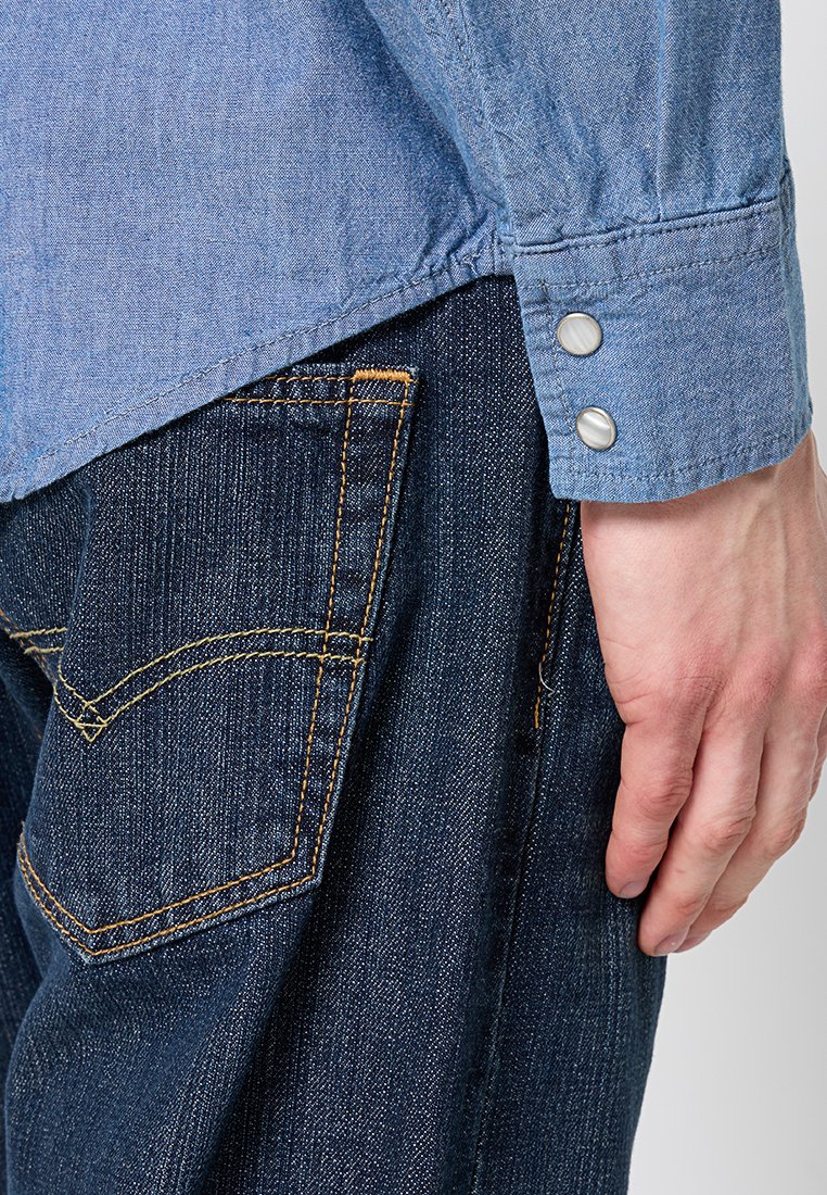 Camisa de mezclilla azul claro de mangas largas, con puños a presión y dobladillo curvado, combinada con jeans azul oscuro que presentan costuras amarillas y bolsillos.