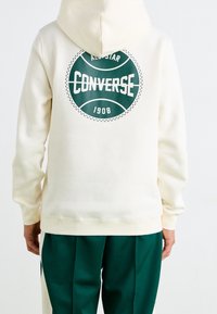 Sweat à capuche de couleur crème avec un grand cercle vert imprimé portant les inscriptions "ALL STAR" et "CONVERSE 1908" au dos. Tissu doux en mélange de coton.