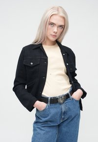 Crna traper jakna s dva prednja džepa, nošena preko bež majice. Modelove svijetloplave traperice imaju dekorativnu kopču za pojas.