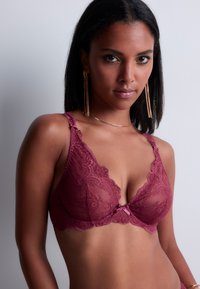 Soutien-gorge en dentelle bordeaux avec des motifs floraux, des bords festonnés, des bretelles ajustables et un petit nœud en satin rose au centre.