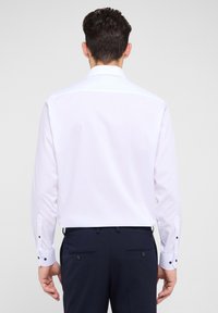 Weißes Langarmhemd mit klassischem Kragen, Knopfmanschetten mit marineblauen Akzenten, kombiniert mit dunkelblauen Hosen. Glatte Stoffoberfläche.