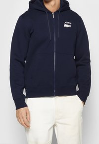 Námořnicky modrá mikina na zip z měkké látky; má klokánkovou kapsu, kapuci se stahovací šňůrkou a bílý logo Lacoste na levé straně hrudi.