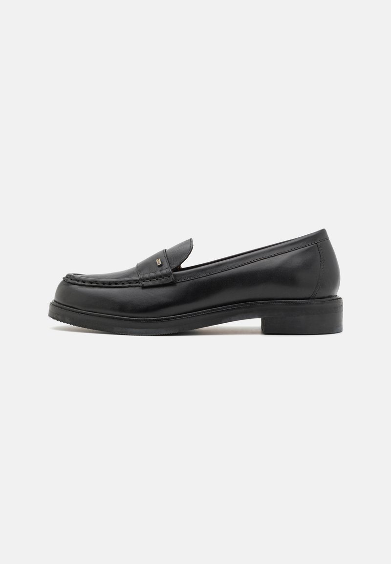 Sapatilhas de couro pretas com bico costurado em estilo mocassino, sola plana e detalhe de hardware discreto na parte superior. Textura suave e design clássico.