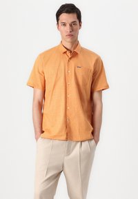 Camisa laranja de manga curta com um acabamento texturizado, fecho de botão e um bolso no peito. Combinada com calças bege de pregas.