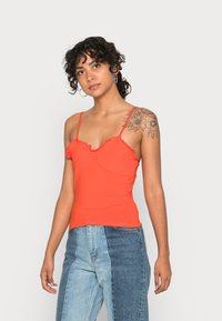 Top de alças ajustado vermelho com bordas em onda, alças finas e uma textura suave. Combinado com jeans de denim com um design em patchwork.