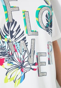 Camiseta blanca con el texto "FLOWER" en letras en negrita y con joyas. El diseño incluye gráficos florales coloridos con acentos en azul, verde y rojo.