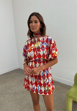 Γυναίκα που φοράει κοντό φόρεμα με κόκκινο και λευκό μοτίβο, μεγάλα σκουλαρίκια κρίκους και κολιέ με μενταγιόν σε σχήμα ήλιου, στέκεται μέσα σε εσωτερικό χώρο κοντά σε μια πράσινη καρέκλα.